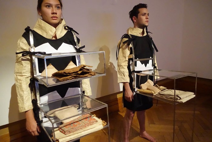 Two people standing side by side in a white gallery space. They wear tan, white and black barkcloth outfits with shelf-like cases containing textile samples.