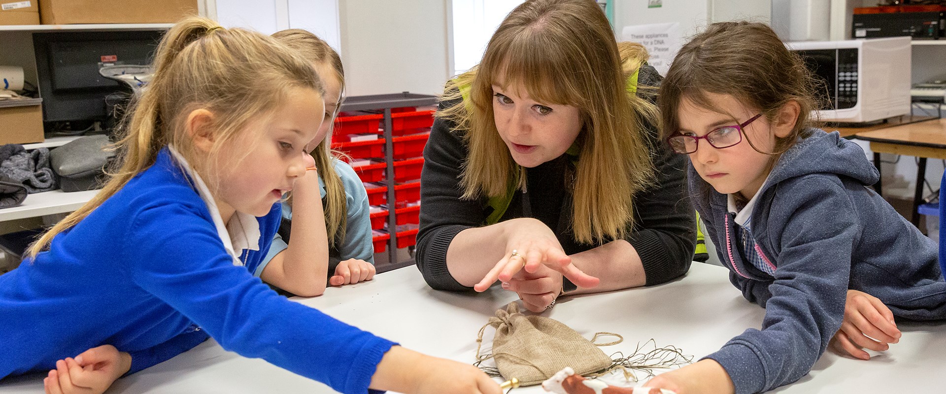 A member of education staff is surrounded by three pupils looking at roman objects on a table. 