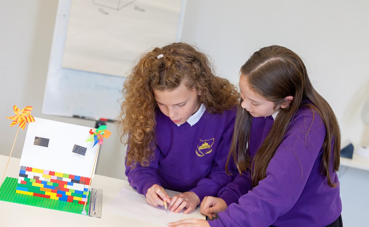 two pupils do some calculations on paper next to a small lego house, which has mini solar panels on the roof and windmills outside. 