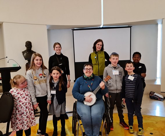 A group of children stand around a person sitting in a wheelchair playing a banjo
