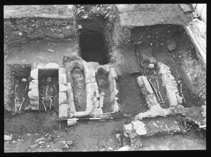 Black and white photo of an excavation pit revealing deep stone foundations, within which are a row of stone cells each containing a skeleton.
