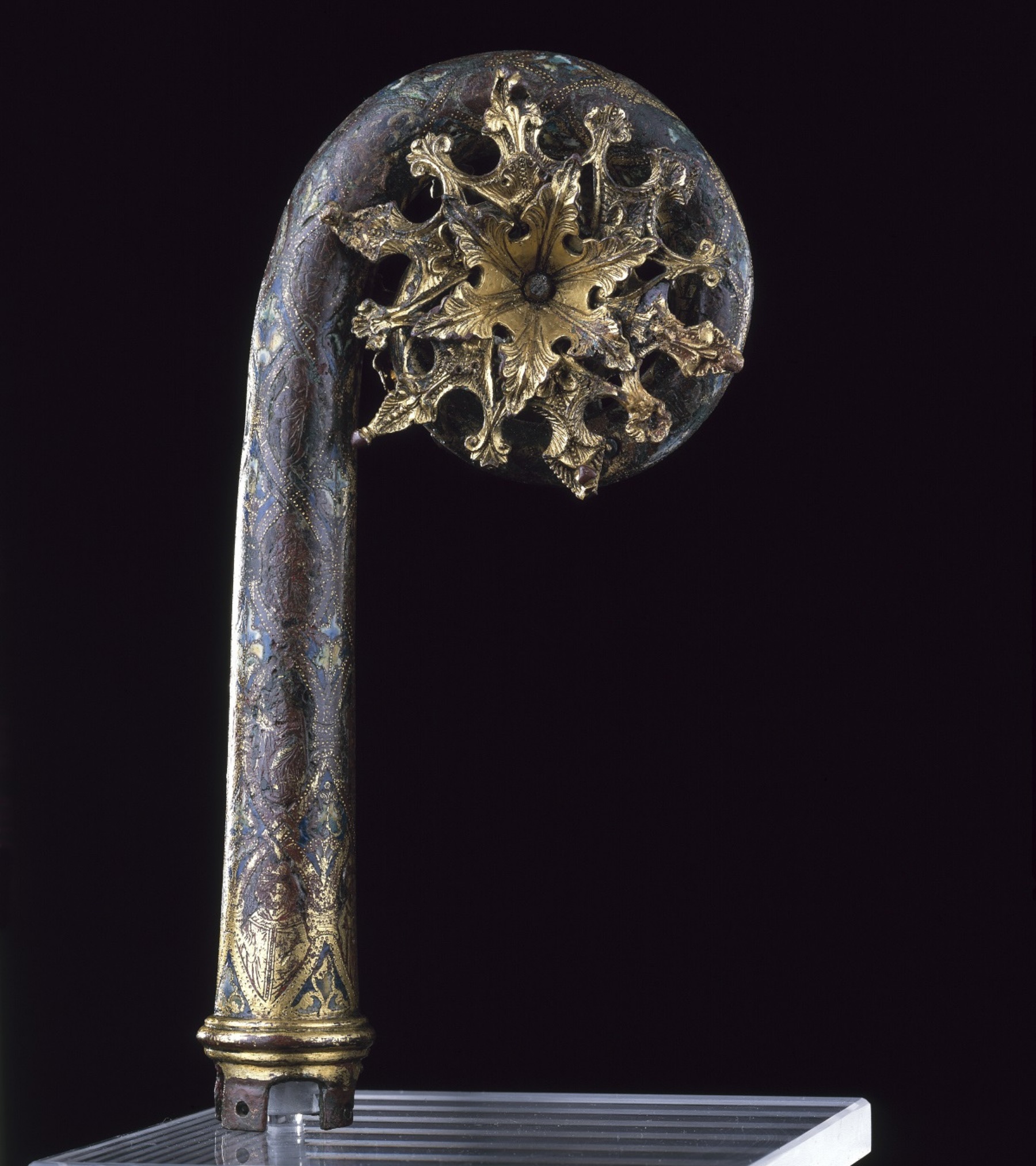 Metalwork object like a cross between a staff and a fiddlehead fern. The staff is grey-blue, and a golden sun-like burst is within its coiled top.