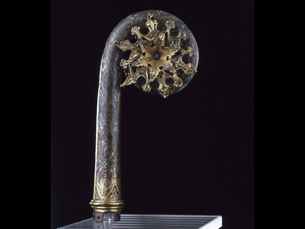 Metalwork object like a cross between a staff and a fiddlehead fern. The staff is grey-blue, and a golden sun-like burst is within its coiled top.