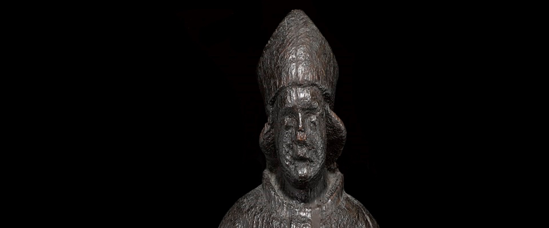 Wooden figure of a saint in flowing robes. The figure is heavily worn and darkened, its arms missing and its face eroded to seem melancholic.