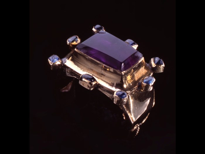 Ornate ring with a large rectangular purple gemstone surrounded by eight smaller purple gemstones mounted on a thick gold ring.