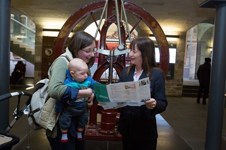 A woman holding a baby looks at a map held by a member of the visitor experience staff.