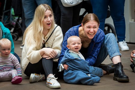 Two women with babies sit on the floor smiling, looking at something beyond the camera.