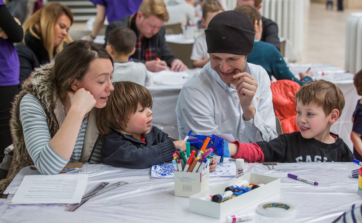 A family with two small children sit at a table with coloured markers, paper, and glue.