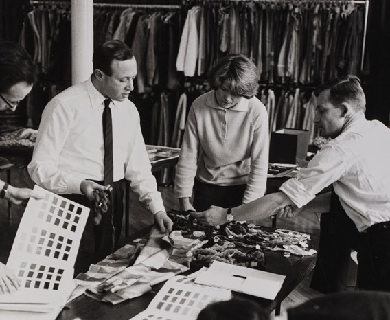 Black and white photo of 4 people standing around a table filled with fabric and yarn samples.