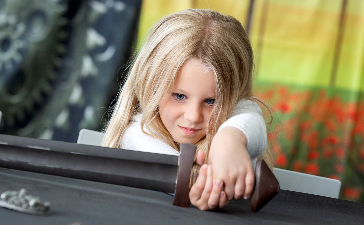 A young child holds the handle of a metal sword as it lies across the table