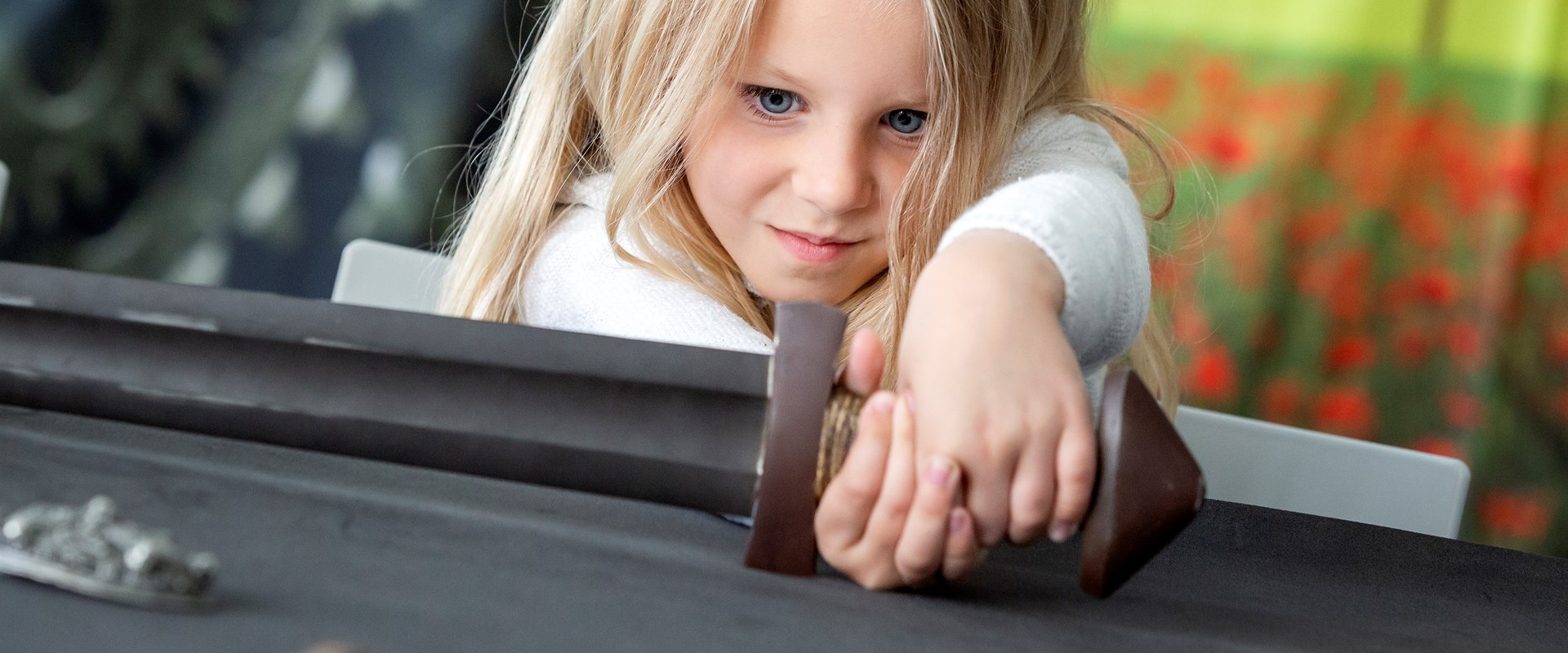 A young child holds the handle of a metal sword as it lies across the table