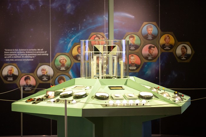 A light green TARDIS console covered in dials and buttons. Behind it is a wall with a galaxy-themed wallpaper and portraits of the Doctor Who actors through the years.