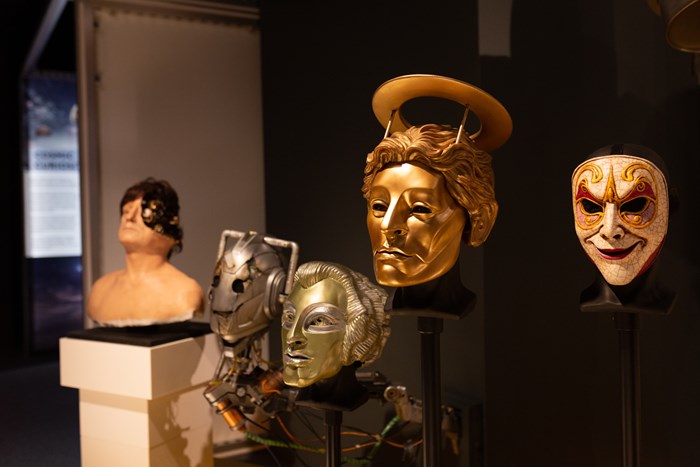 Five |Doctor Who monster masks mounted on poles in a museum gallery. Masks include Cybermen, Heavenly Host, and the droids from 'The Girl in the Fireplace'.