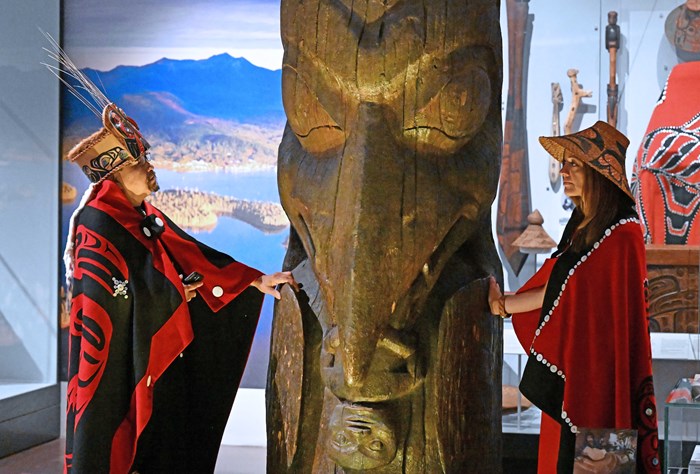 Two people in ats and red and black robes stand on either side of a large carved wooden pole.