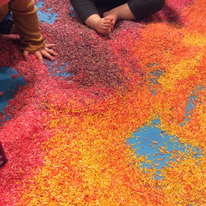 A blue floor covered in dry rice grains dyed in orange, red and pink. The hands and feet of two small children are seen at the edge of the frame.