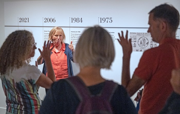 A woman stands in front of a printed timeline on a wall signing in BSL. In the foreground, out of focus, 3 visitors face her, two with hands raised.