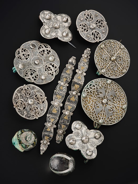 Array of eleven metal objects on a black background. Two decorated silver and gold arm-rings are in the middle, surrounded by disc brooches, quatrefoil brooches, and small crystal spheres with metal casings.