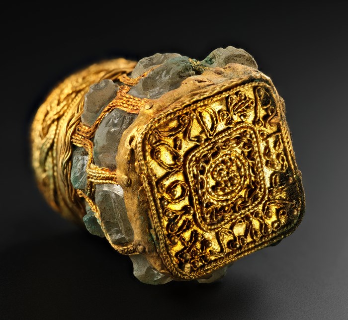 A green and vivid gold rock crystal jar laid on its side with the bottom facing the camera. Loopy Latin letters form a square around a densely decorated centre, the patterns resembling rope or beads.