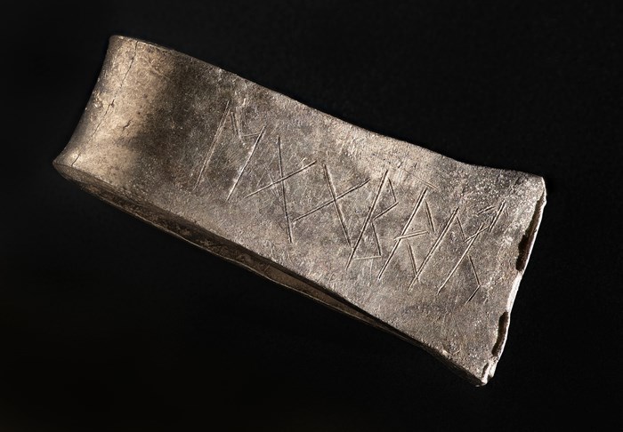 A wide flat silver arm ring viewed from the side. It has Anglo-Saxon runes carved into it.