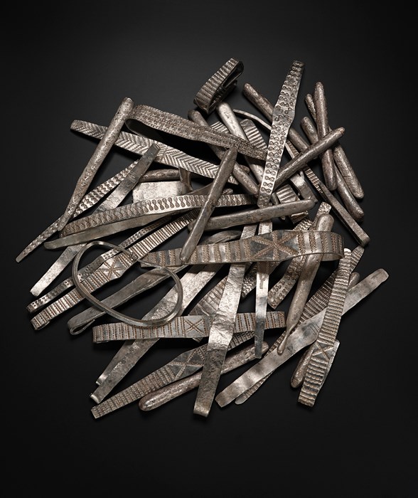 Pile of several dozen silver arm-rings on a black surface. The arm-rings are elongated rectangles, many decorated with simple lines, x-shapes, chevrons, and small circles.