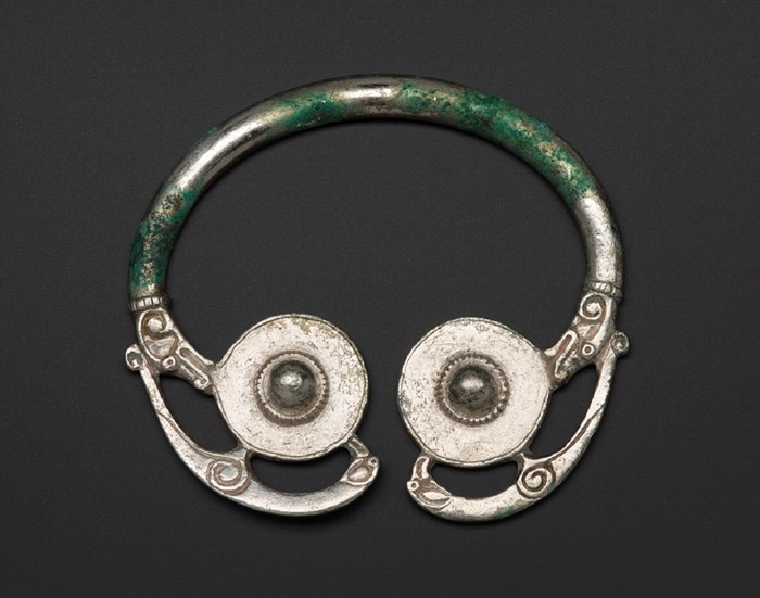 Silver brooch on a dark grey background. The brooch has two shield-like discs at the bottom at each end. Thin silver birds branch off from the discs and into the thin upper hoop.