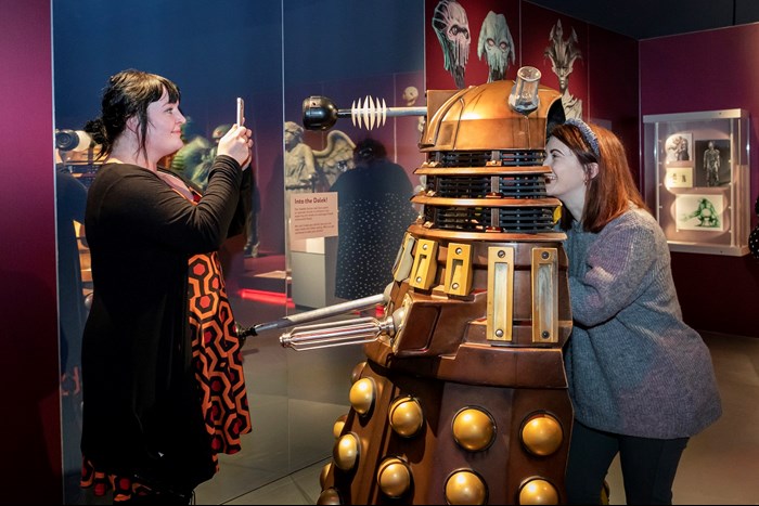Inside the Doctor Who exhibition, a woman in a grey jumper steps inside a Dalek costume and another woman in a black cardigan takes a picture.