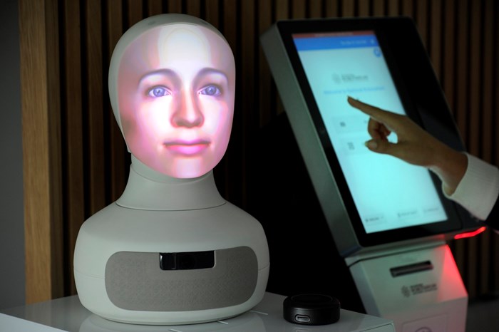 A human face is digitally overlaid on a white robotic head-shaped stand. A screen controls the face's actions, with a person's hand hovering over it.