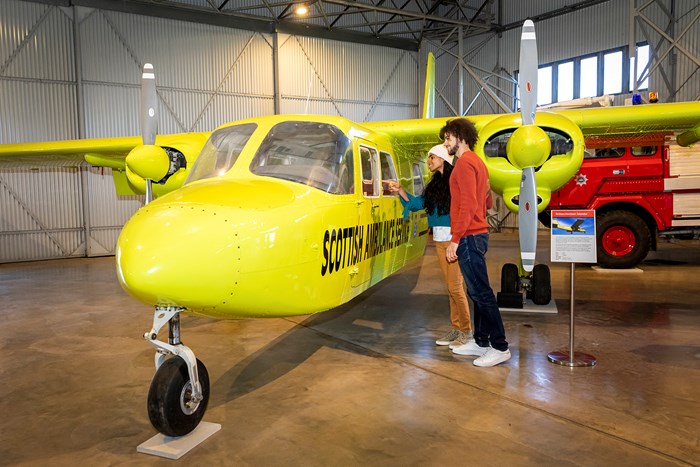 Two visitors looking inside the cockpit of a yellow aircraft parked in a storage hangar.