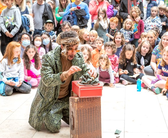 dozens of children sat around a dressed up storyteller in the centre. All people look amazed and enchanted.