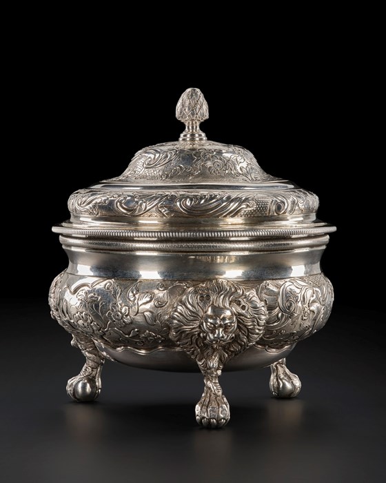 A shiny silver vessel, squate and wide with three legs made to look like lion's paws, engravings of lions with wide, circular manes alongside simple flowers on vines, and a heavy lid.