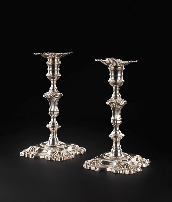 A pair of silver candlesticks against a black surface. The silver is exceptionally shiny and fine. Each candlestick has a thick, square base, a thin square platform at the top, and three flared sections on the stick.