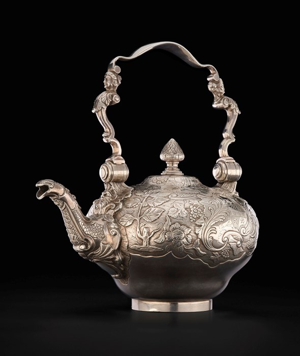 A very ornate silver tea kettle against a black background. The spout has scales almost like a fish, the handle is very delicate with an aristocratic figure on each side, and the body of the kettle is covered in engravings of trees and flora.