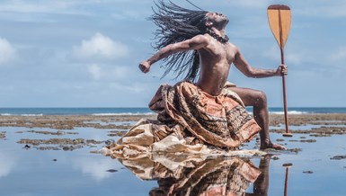 A man from Oceania dramatically poses on a beach covered in a shallow layer of water and seaweed. Throwing his head back and striking a 'warrior pose', his long braided hair, traditional dyed cloth wrapping below his waist, and the boat paddle he holds make him seem powerful and heroic.