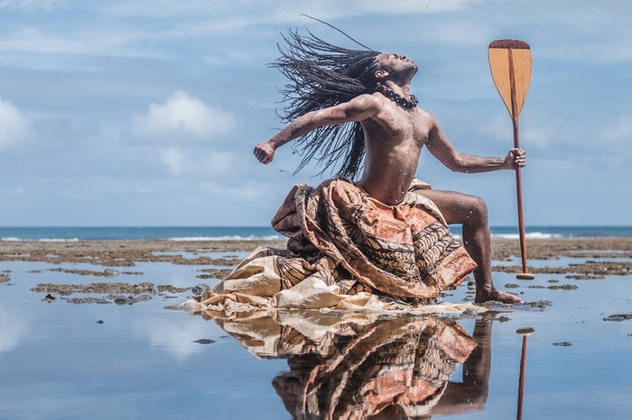 A Pacific Islander man dramatically poses on a beach covered in a shallow layer of water and seaweed. Throwing his head back and striking a 'warrior pose', his long braided hair, traditional dyed cloth wrapping below his waist, and the boat paddle he holds make him seem powerful and heroic.