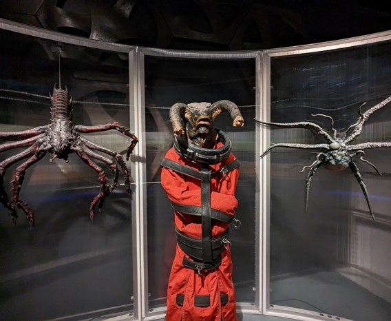 A dark museum gallery space with metal and mesh panels reminiscent of a sci-fi setting. A large central figure is a humanoid alien with two trunk-like appendages on its head wrapped in an orange straightjacket. It is flanked by two Daleks clinging to the wall, resembling a mix of spider, lobster, and insect.