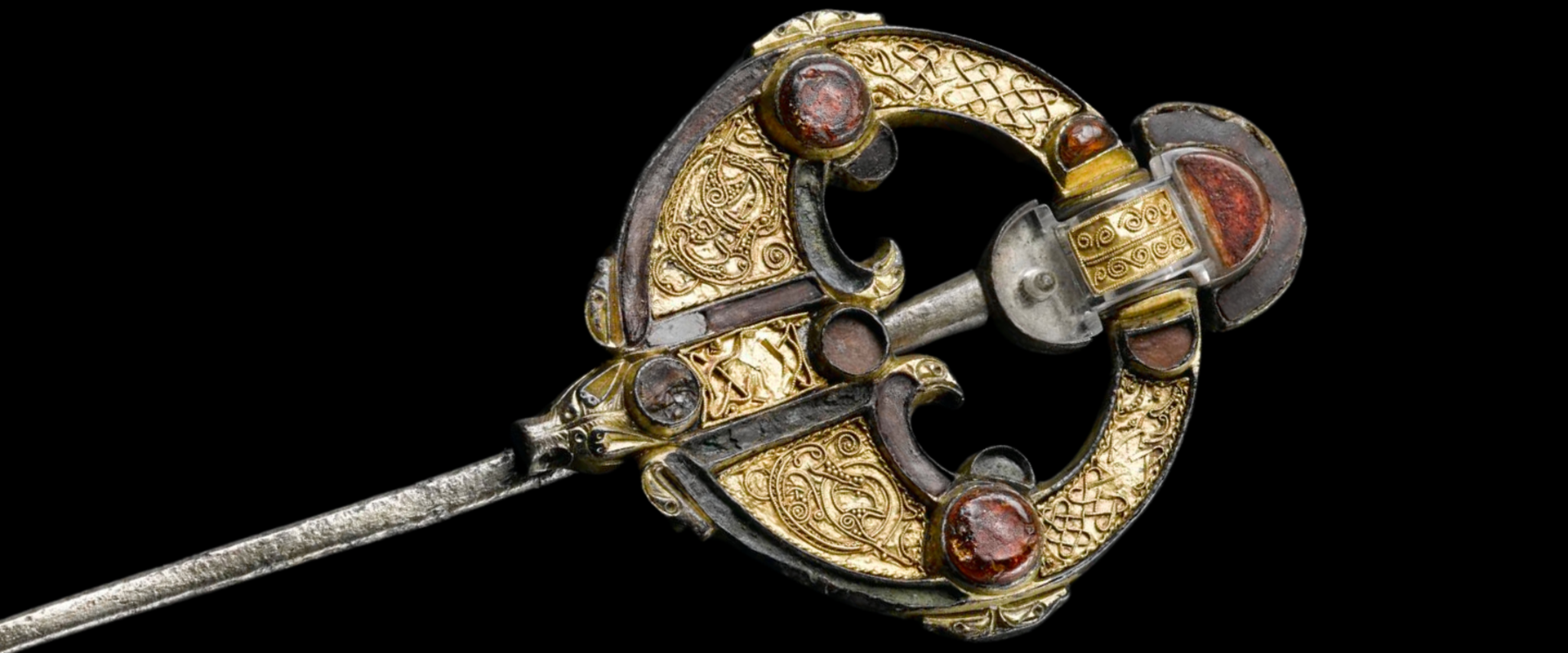 Gold-coloured brooch and pin, intricately decorated with raised Pictish-style patterns and red gemstones.