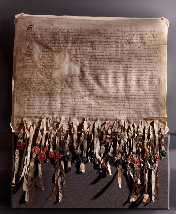 The Declaration of Arbroath laid out on a grey protective base. The Declaration has dense Latin script across it with two small patches missing, and many strips dangling from the bottom affixed with green and red wax seals.