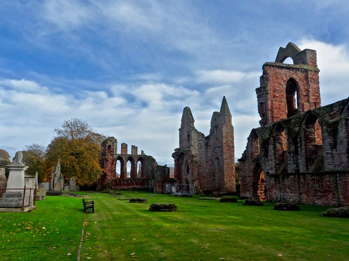 A large, ruinous medieval abbey made from red sandstone. A tall, hollowed-out tower and stretches of walling flank a green grass field with several graves in it.
