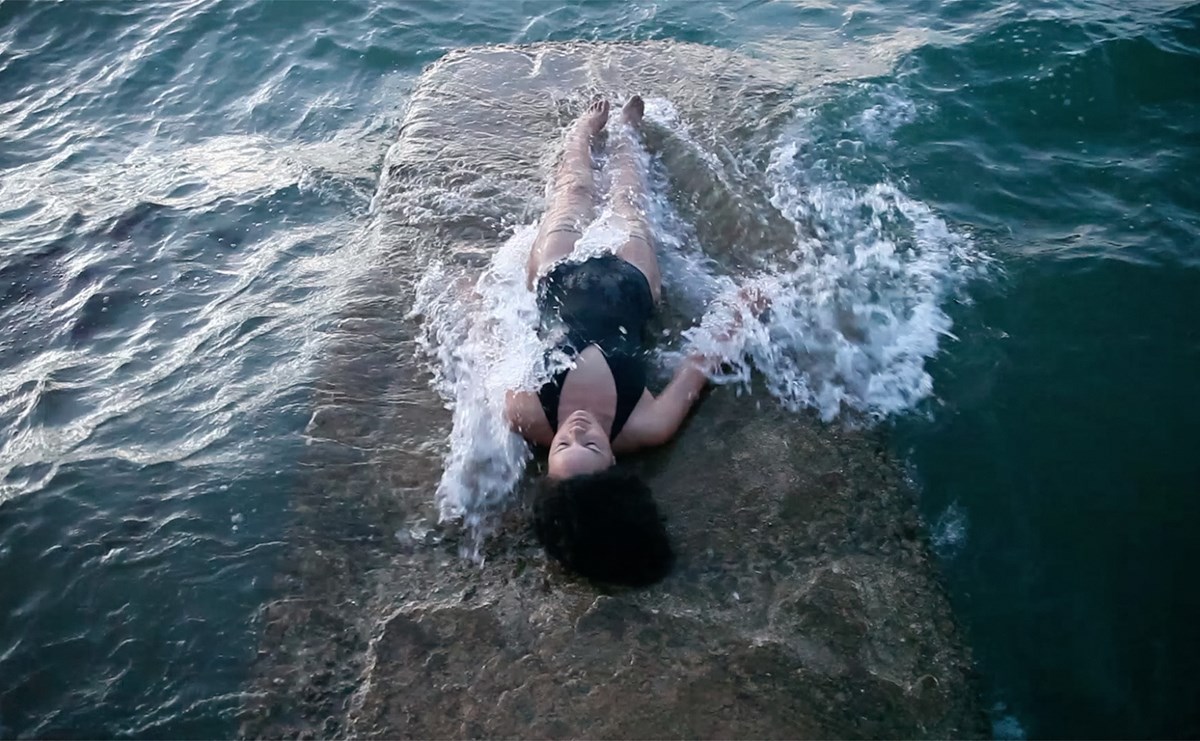 A woman lies facing up in the surf on a beach, with a small wave washing over her.