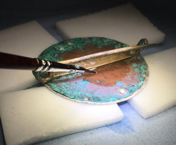 The circular brooch rests atop four rectangular pads under a bright light. A metal tool is being used to delicately remove corrosion from its copper and green surface.