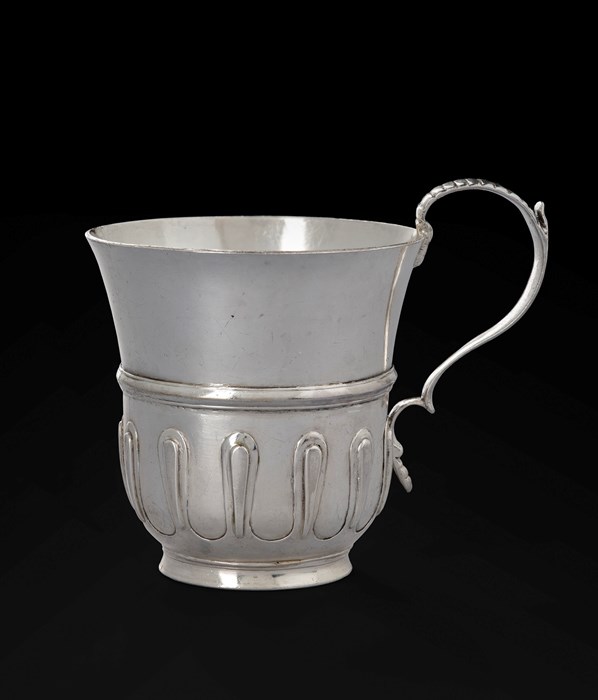 A tall, hefty silver vessel resembling a jug against a black background. Its top widens to resemble a thistle. It has a very spindly handle and a simple, looping engraving pattern near the bottom.