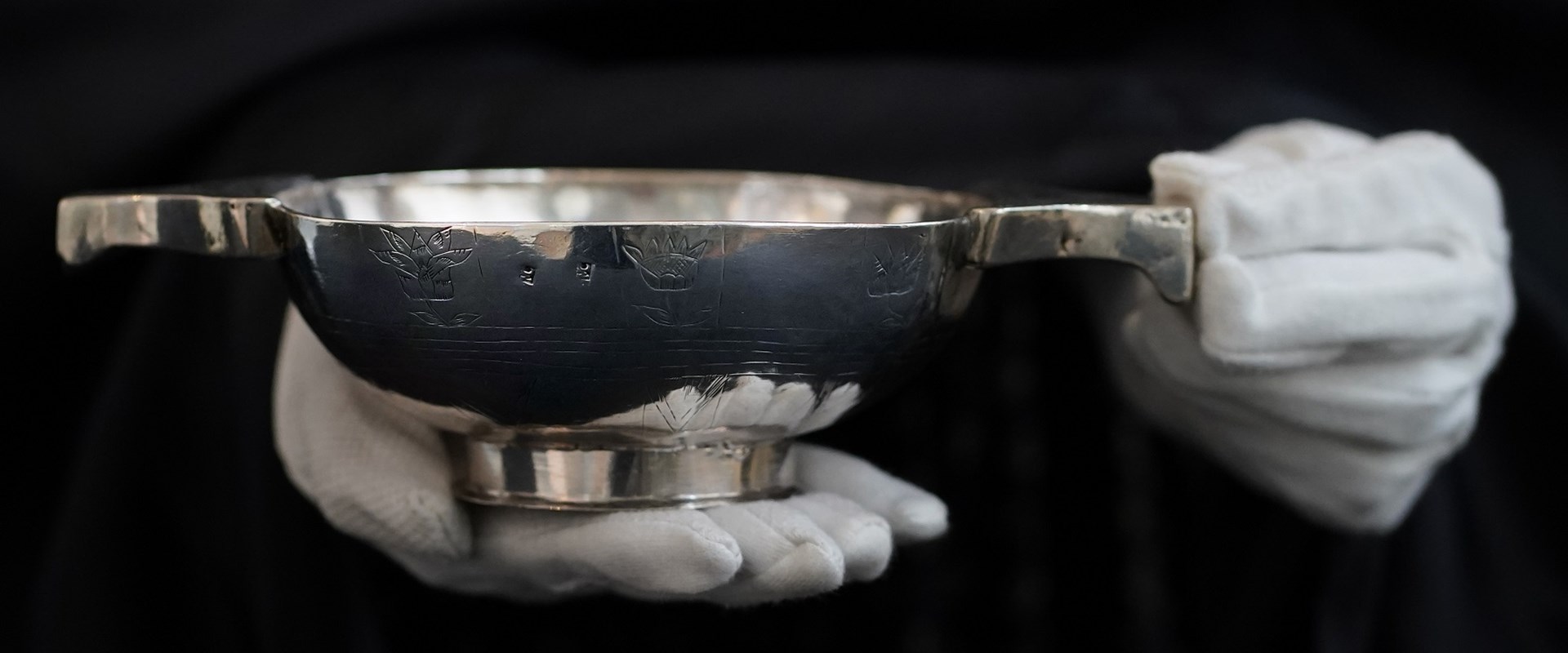 A shiny silver quaich being held delicately by two white-gloved hands against a black background.