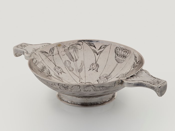 A silver quaich viewed at a high, oblique angle against a light grey background. The inside and outer surface of the quaich are covered in floral engravings.