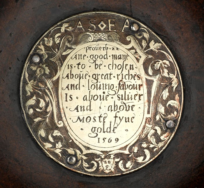 Closeup of a gold-coloured engraved plate in the mazer's wooden bowl. Words in a stylised script sit above the year '1569'.