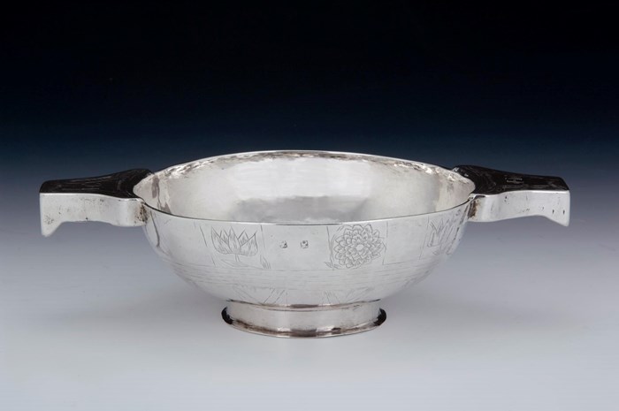 A shiny silver quaich, or shallow dish, with two flowers engraved on its surface and two short, stout arms branching out from the rim.