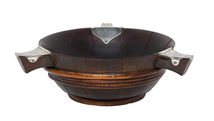 A wooden quaich, or shallow dish, which looks as though it could be made from a whisky barrel. It has three short, stubby arms branching out from the rim, each with a small silver covering.
