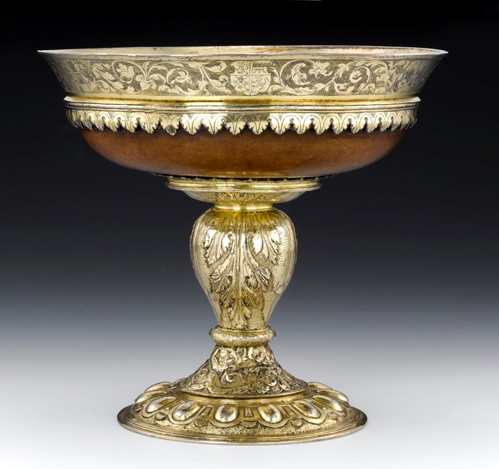 A mazer, a type of cup, viewed from the front against a grey and black background. The mazer's bowl is made of wood, lined with gold-coloured silver covered in engraved floral designs. The mazer's stem is entirely metal with more floral engravings.