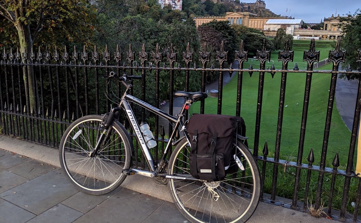 A black and silver hybrid bicycle laden with pannier bags rests against a black iron railing in Edinburgh, with the National Gallery and Princes Street Gardens in the background.