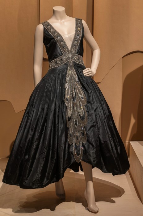 Female mannequin wears a black dress with a deep v-neck, a wide skirt, and beaded detail along the neckline and down the front.
