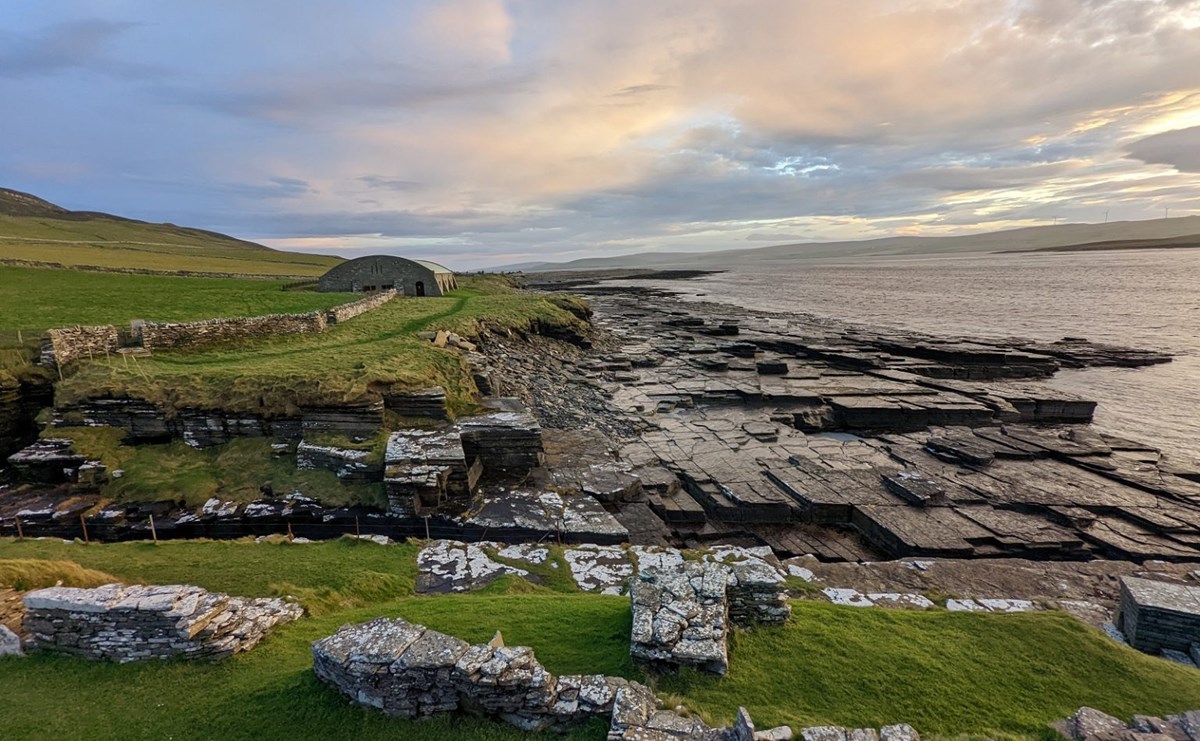Under broad, cloudy skies turning colourful at sunset is a stony shoreline with several stone structures built near the edge. Rectangular flagstones slope down into the waters, and a rock trench cuts of a promontory in the foreground. A faint path leads to a hangar-like structure in the middle distance.